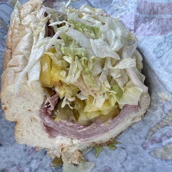 Wawa - Italian Hoagie - A sandwich with sliced ham, shredded lettuce, and pickles on a soft, white bun. The sandwich is partially wrapped in paper, placed on a surface with a car interior visible in the background. The ingredients are layered, with the lettuce and pickles on top of the ham, inside the bun. - Gas Stations Near Me - Sterling, Virginia