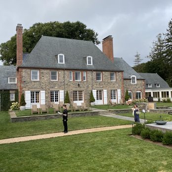 Shepherd’s Run - A large stone estate with a steep roof, multiple chimneys, and white window shutters, surrounded by a manicured lawn and outdoor seating. - Venues & Event Spaces Near Me - South Kingstown, Rhode Island