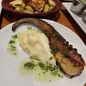 La Malinche - Pulpo - A white plate holds a grilled octopus tentacle topped with chopped herbs, accompanied by a serving of mashed potatoes. The dish is garnished with green herbs and oil. In the background, there are additional plates with food, including a piece of cooked meat and vegetables, on a wooden table. - Tapas Bars Near Me - Silver Spring, Maryland