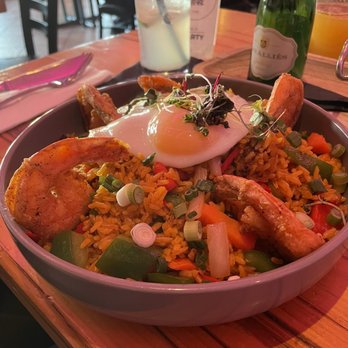 Travesias - Shrimp wok - A bowl of fried rice with vegetables, topped with a poached egg and microgreens, served with fried plantains, on a wooden table in a restaurant. - Latin American Near Me - Bronx, New York