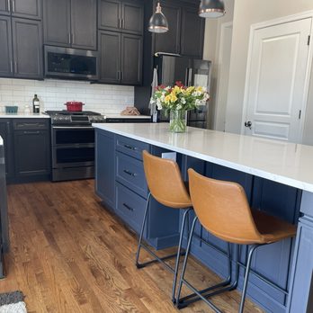 Cabinet Tree Studio - The image shows a contemporary kitchen with dark gray upper cabinets and a matching microwave. The kitchen has a white subway tile backsplash and a stainless steel stove. A white countertop island with a blue base includes drawers and cabinets, with a glass vase of flowers placed on top. Two wooden bar stools with metal legs are positioned at the island. The flooring is hardwood, and there is a white door on the right side of the image. - Interior Design Near Me - Plainfield, Illinois