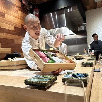 Hiden - A sushi chef, dressed in traditional white attire, presents a wooden box filled with sashimi, including slices of tuna and other raw fish, to diners seated at a wooden counter. The restaurant features a wooden wall and a stainless steel kitchen hood in the background. Diners have glasses of wine and are engaged in conversation, with a modern Asian art piece on the wall. - Japanese Near Me - Miami, Florida