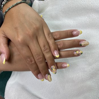 Charming Nails - Simple - The image shows a person's hands with freshly manicured nails, decorated with yellow tips and floral patterns. The hands are resting on a white towel, and the background includes various nail polish bottles and salon supplies. The person is wearing multiple bracelets on their wrist, and the nails are well-groomed with a glossy finish. - Nail Salons Near Me - Washington, DC, District of Columbia