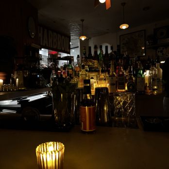 The Snail - A bar interior features a counter with numerous bottles of liquor illuminated from behind, creating a warm, glowing effect. The ceiling has small pendant lights, and a candle on the table adds to the ambient lighting. The background includes a sign with the word 'HARBOR' and some framed artwork on the wall, suggesting a cozy, vintage atmosphere. - American Near Me - Brooklyn, New York