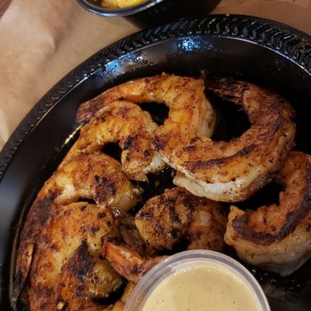 Mission BBQ - Specialty shrimp dish - The image shows a black plastic tray containing grilled shrimp with a golden-brown, slightly charred exterior, accompanied by a small cup of creamy, yellow dipping sauce. In the background, there is a bowl of macaroni and cheese with a cheesy, yellow sauce. The setting appears to be a casual dining environment, with a wooden table surface visible. - Barbeque Near Me - Springfield, Illinois