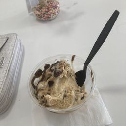 Salted Caramel Brownie - A cup of vanilla ice cream topped with chocolate syrup and mix-ins is placed on a white table with a black spoon.