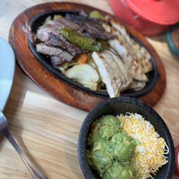 Combo Fajita Platter - The image shows a sizzling platter of grilled meats, including beef and chicken, accompanied by grilled vegetables such as peppers and onions, served on a wooden tray. A small black bowl contains guacamole topped with shredded cheese. The setting appears to be a casual dining environment with a wooden table and various containers in the background.