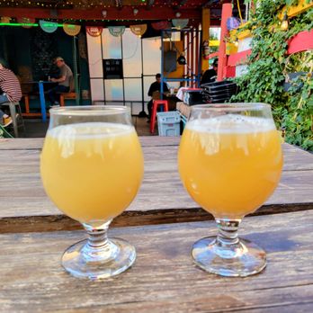 Finn's Manor - Two glasses of pale yellow beverage are placed on a wooden table at an outdoor bar with colorful string lights and patrons in the background. - Cocktail Bars Near Me - Denver, Colorado