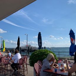Come for the view if nothing else - Seaside restaurant patio with tables, chairs, umbrellas, and patrons dining near the water under a blue sky.