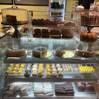 DC Vegan - Bakery - A bakery display case contains a variety of vegan baked goods, including cakes, cookies, cupcakes, and macarons, arranged on multiple shelves. The top shelf features cakes on stands, with one decorated with chocolate frosting and another with a pumpkin decoration. The middle shelf displays slices of cake and whole cookies, while the bottom shelf holds assorted cookies and macarons in different colors. Decorative pumpkins and plant-themed elements are placed around the display, indicating a seasonal or autumn theme. - Vegan Near Me - Washington, DC, District of Columbia
