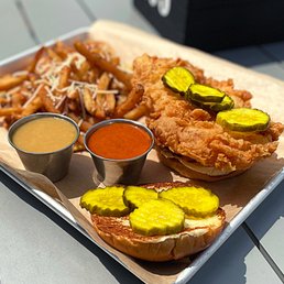 Cluck Yeah Chicken Sandwich - A fried chicken sandwich topped with pickles, served with seasoned fries and two small cups of dipping sauces, on a tray.