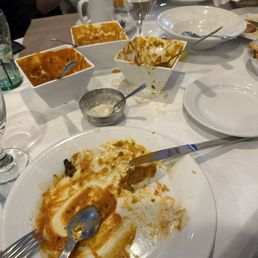Demolished... - A table with multiple used dishes, bowls, and utensils, including empty bowls with remnants of sauce, a large plate with leftover food, and various utensils. There are glasses of water and wine, a coffee mug, and a small bowl with sauce. The setting suggests a restaurant after a meal, with some food and drink remnants visible.