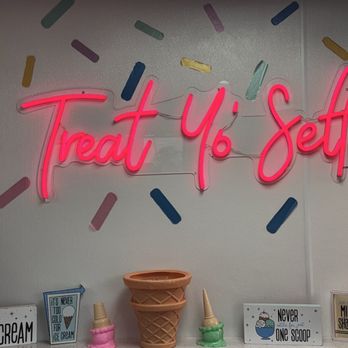 JJ's Custard Co - A neon sign with the phrase 'Treat Yo Self' in pink is mounted on a wall decorated with multicolored sprinkles. Below the sign, there are several ice cream-themed signs and small decorative items, including ice cream cone models and signs with phrases like 'Ice Cream' and 'Never eat just one scoop.' The setting appears to be an ice cream shop or dessert-themed area. - Ice Cream & Frozen Yogurt Near Me - Dearborn, Michigan
