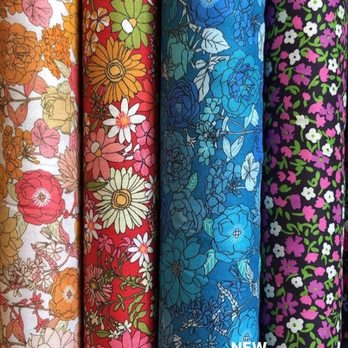 Harmony - Four rolls of fabric are displayed vertically, each featuring a floral pattern. The first has orange, pink, and beige flowers on a white background. The second features red, green, and white flowers on a red background. The third showcases blue flowers on a blue background. The fourth has small white, purple, and pink flowers on a black background. The image includes text advertising new cotton lawns at $12 per yard. - Fabric Stores Near Me - Provo, Utah