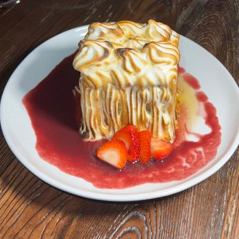 Platea - Baked Alaska - A baked Alaska dessert with toasted meringue, strawberry slices, and berry sauce on a white plate, served on a wooden table. - Peruvian Near Me - Pinecrest, Florida