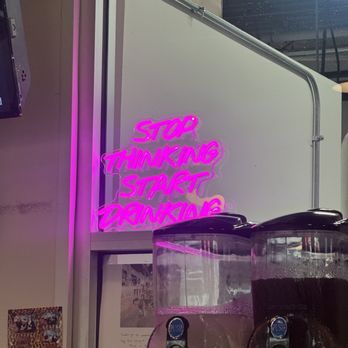 Thick-N-Thin Brewing - Pink neon sign reading "Stop Thinking Start Drinking" is mounted on a white wall inside a brewery or bar. In front of the sign, there are beverage dispensers with clear containers, one filled with a light-colored liquid and the other with a darker liquid. The setting includes visible interior elements such as a window, some wall decorations, and part of the ceiling, indicating an indoor social venue focused on drinks. - Breweries Near Me - Hagerstown, Maryland