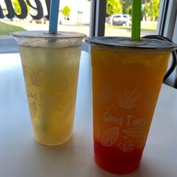 Mango Green Tea - Two beverages in clear cups with straws are placed on a white table near a window, with a view of trees and parked cars outside.