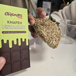 Snickers Chocolate Bar - The image shows a person holding a chocolate bar labeled 'KNAFEH PISTACHIO' with Belgian milk chocolate, and another hand holds a piece of chocolate covered with sprinkles. The background includes a table with a clear plastic container and a bag, indicating an indoor setting, possibly a dessert shop or cafe.
