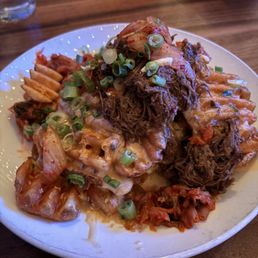 A white plate holds a Korean-style meal with shredded beef, kimchi, and various side dishes. The shredded beef is topped with chopped green onions, and the kimchi is visible around the edges. The meal is served on a wooden table with a glass of water and a stack of plates in the background.