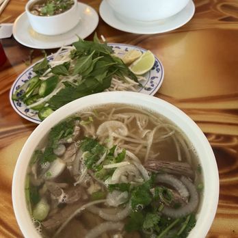 Pho 14 - A bowl of Vietnamese pho with sliced onions, green herbs, and beef, accompanied by lime wedges and fresh herbs on a plate. - Vietnamese Near Me - Washington, DC, District of Columbia