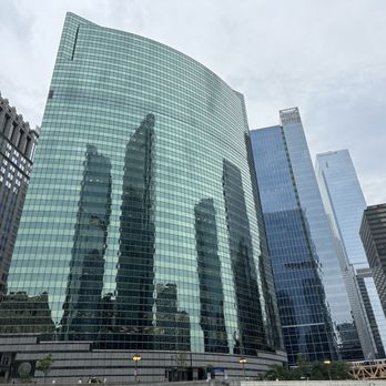 Chicago Architecture Center - A large, curved glass skyscraper dominates the scene, reflecting the images of surrounding high-rise buildings in its facade. The building is part of a dense urban area with multiple modern office towers, some with blue-tinted glass and others with darker exteriors. The sky above is overcast, and the base of the building features a small plaza with street lamps and a few trees. - Historical Tours Near Me - Chicago, Illinois