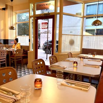 Buttercup  - The image shows the interior of a restaurant with multiple tables arranged for dining, each set with napkins, glasses, and small candles. Large windows allow natural light to enter, and a woman is seated at a table near the window on the left side. The decor includes pendant lighting and a patterned floor. The entrance door is visible, with an exit sign above it, and outside, parked cars can be seen through the windows. - Coffee & Tea Near Me - Chicago, Illinois