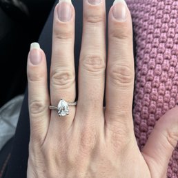 A close-up of a hand wearing a diamond engagement ring with a pear-shaped diamond, resting on a person's lap with a pink knitted fabric nearby.