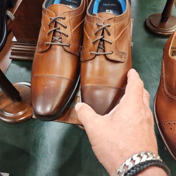Dillard's - A pair of brown leather dress shoes with laces are positioned on a wooden display surface in a retail store. The shoes have a polished appearance with a darker toe cap and are lined with a teal-colored interior. A person's hand, wearing a silver bracelet and a black braided bracelet, is holding one of the shoes. The background includes other shoes and furniture, indicating a shoe store environment. - Department Stores Near Me - Plantation, Florida