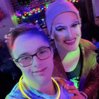 The Bistro - a man and a woman posing for a picture - A selfie features a person with glasses, wearing a tie-dye shirt and a glowing yellow necklace, standing next to a drag performer with dramatic makeup, a beanie, and a leather jacket. The background shows a brick wall decorated with wreaths and colorful string lights, indicating a festive or holiday event at a venue associated with a gas company. - Gay Bars Near Me - Bloomington, Illinois