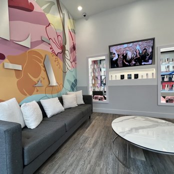LaserAway - Lobby - A modern waiting room features a large floral mural on one wall, a gray sofa with white pillows, and a wall-mounted TV displaying a group photo. - Tattoo Removal Near Me - Washington, DC, District of Columbia