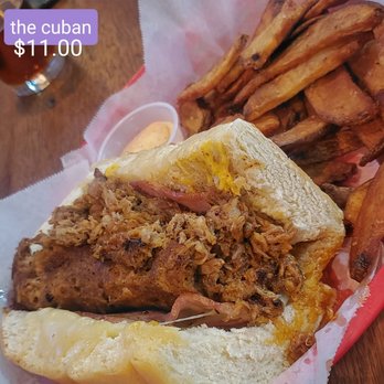 DT Kirby's - Crazy fat in all the ways! Stupid upcharge for fries. I prefer the Reuben at Town & Gown and The Tap burgers. - A Cuban sandwich featuring layers of pulled pork, ham, melted cheese, and pickles inside a toasted bread roll, accompanied by a side of French fries and a small container of dipping sauce. The meal is presented on a red tray lined with paper, with a drink visible in the background. - Burgers Near Me - Lafayette, Indiana