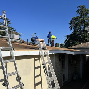 Steep Flat Roofing - Three workers are on the roof of a residential house, engaged in roofing work. One worker is kneeling, possibly securing materials, while the other two stand nearby, observing or assisting. Two ladders are positioned against the house, providing access to the roof. The house has a beige exterior with a small porch area, and the sky is clear and blue, indicating good weather for construction work. - Roofing Near Me - Yucaipa, California