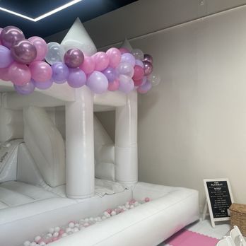 The Loft - An inflatable white castle structure decorated with clusters of pink, purple, and white balloons along the top edge. The castle has towers and a small entrance, with a ball pit filled with pink, white, and purple balls at the base. A small blackboard sign and a wicker basket are positioned nearby, with chairs partially visible in the foreground. The setting appears to be an indoor event space or party area. - Venues & Event Spaces Near Me - Long Beach, California