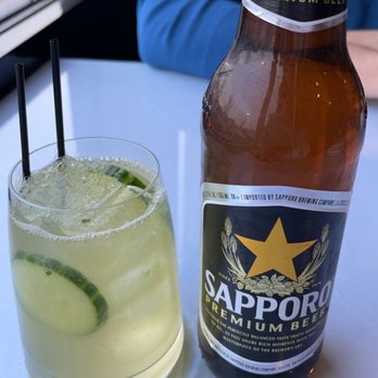 Thai Chada - Drinks - A Sapporo beer bottle and a cucumber cocktail with ice and cucumber slices are placed on a white table, with a person in a blue shirt in the background. - Cocktail Bars Near Me - Bellingham, Massachusetts