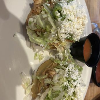 Los Cantaros - Two tacos with shredded lettuce, cheese, and sauce are presented on a white plate, accompanied by a small black cup of sauce or soup. - Mexican Near Me - Hampton, New Hampshire