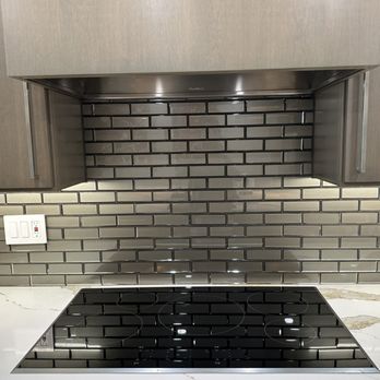 First Stop Flooring - Kitchen backsplash - The image shows a kitchen countertop featuring a black electric cooktop with four burners. Behind the cooktop, there is a gray subway tile backsplash extending to the upper cabinets. The cabinetry above is gray with metallic handles, and a white marble countertop with subtle veining is visible on the left and right edges of the image. An electrical outlet is mounted on the backsplash to the left of the cooktop. - Flooring Near Me - Phoenix, Arizona