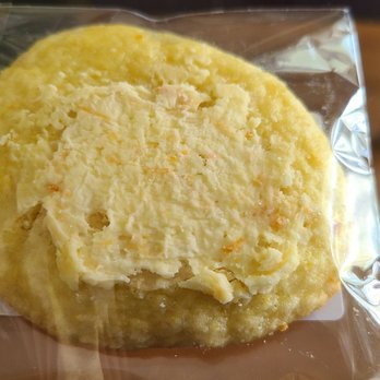 Bake Share Baking - Orange drop cookie - A single orange drop cookie is sealed in clear plastic packaging, revealing its round shape and crumbly texture. The cookie has a slightly uneven surface with visible small cracks and a light orange hue, indicating a citrus flavor. It is placed on a flat surface, with a small piece of paper or receipt partially visible underneath the packaging. - Desserts Near Me - Grayslake, Illinois