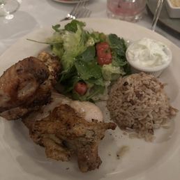Half Chicken - A meal plate includes grilled chicken pieces, a portion of rice, a fresh salad with lettuce, tomatoes, and greens, and a small bowl of yogurt or tzatziki sauce. The dish is served on a white plate with a table setting that includes glasses and utensils, indicating a dining experience at a Greek restaurant.