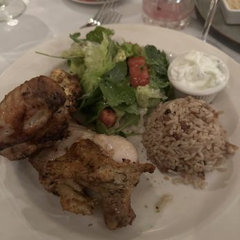 Athos - Half Chicken - A meal plate includes grilled chicken pieces, a portion of rice, a fresh salad with lettuce, tomatoes, and greens, and a small bowl of yogurt or tzatziki sauce. The dish is served on a white plate with a table setting that includes glasses and utensils, indicating a dining experience at a Greek restaurant. - Greek Near Me - Albany, New York