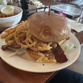 The Joint - Stony Brook - A burger with a toasted bun, fried onion rings, bacon, lettuce, and sauce is served on a white plate. Accompanying the burger are French fries and a bowl of fried chicken with a side of dipping sauce. The setting appears to be a casual restaurant with condiments and utensils visible on the table. - Beer, Wine & Spirits Near Me - Boston, Massachusetts
