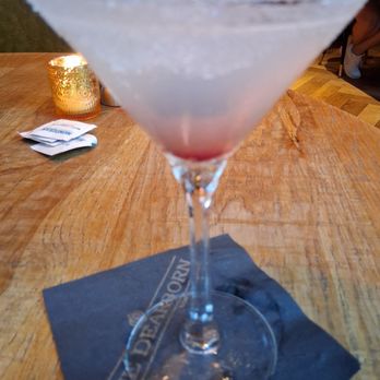 The Dearborn - Lemondrop Martini!! Delicious! - A lemon drop martini with a sugared rim is presented in a clear cocktail glass on a wooden table at The Dearborn restaurant in Chicago. The drink appears to have a pinkish hue at the bottom, with a cloudy, frosted appearance. The glass is placed on a dark napkin with the restaurant's name, and a small candle in a gold holder is visible in the background. The setting suggests a casual dining environment with ambient lighting. - New American Near Me - Chicago, Illinois
