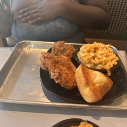 Fried chicken, mac and cheese, and cornbread. - The image shows a metal tray with fried chicken, macaroni and cheese, and a piece of cornbread, served at Cornbread Brooklyn. A person is visible in the background, holding a phone, and a smartphone is placed on the table. The setting appears to be a casual dining environment with a person wearing a patterned top.