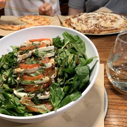 Caprese Salad - A Caprese salad featuring sliced tomatoes, fresh mozzarella, basil leaves, and balsamic glaze is presented in a white bowl on a wooden table. The salad is arranged with layers of ingredients and garnished with herbs. In the background, there are two pizzas on parchment paper, a glass of water, and other beverages, indicating a casual dining setting at Your Pie Pizza in Thornton, CO.
