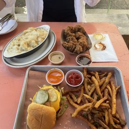 Chicken Nuggets - A meal is displayed on a pink table, including a tray of French fries, a cheeseburger with pickles, a dish of fried chicken, and several small containers of dipping sauces. The setting appears to be an outdoor dining area, with a person partially visible in the background wearing a white shirt and black top. The food presentation includes a variety of condiments and a casual dining atmosphere.
