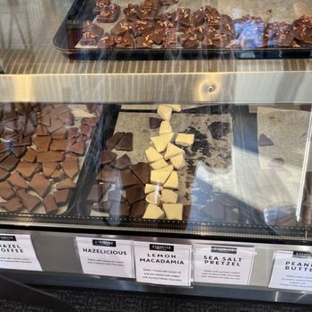 Terry's Toffee - The image features a glass display case containing multiple trays of handcrafted toffee in different flavors, such as Mazel Toffee, Hazlicious, Lemon Macadamia, Sea Salt Pretzel, and Peanut Butter. Each flavor is labeled with a sign, and the toffee pieces are arranged on trays with parchment paper. The case is part of Terry's Toffee, a business known for small-batch confections made with high-quality ingredients, located in Highland Park, IL. - Candy Stores Near Me - highland park, Illinois