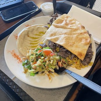 Damashkey Cuisine - Beef Shwarma plate - A plate of beef shawarma served with hummus, a side salad, and pita bread, accompanied by a small container of sauce, on a tray at a restaurant. - Mediterranean Near Me - Lindenhurst, Illinois