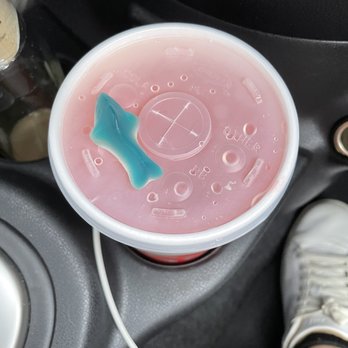 Swig - Unlucky Ducky (Sprite, lemonade, strawberry drink) featuring a shark gummy - A pink beverage in a clear plastic cup features a shark-shaped gummy on top. The cup is situated in a car's cup holder, with part of a person's foot in a white sneaker visible nearby. The car interior includes a black dashboard and a white charging cable. - Desserts Near Me - Gilbert, Arizona