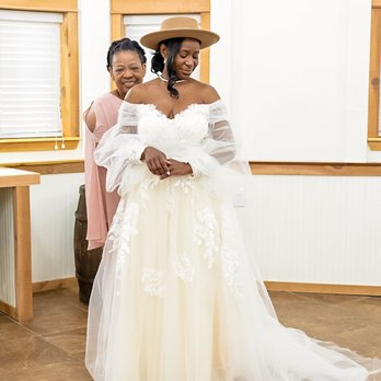 Eivan's Photo Inc - A bride in a white off-shoulder wedding gown with lace details and a beige hat stands indoors, smiling with her eyes closed. Behind her, a woman in a pink dress is smiling and hugging her from behind. - Photographers Near Me - Mokena, Illinois