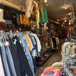 Fun!! - The image shows the interior of a vintage thrift store with clothing racks filled with jeans, shirts, and other apparel. Decorative items such as a feathered swan, a gold mannequin, and hanging dresses are suspended from the ceiling. Shelves display vintage boots and accessories, and the store has a patterned ceiling with multiple light fixtures. The space appears crowded with various vintage and secondhand items arranged throughout.
