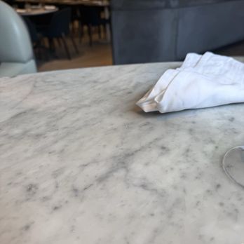 Vela - A marble table with a white cloth napkin on it, situated in a restaurant with chairs and tables in the background. - Venues & Event Spaces Near Me - Toronto, Ontario